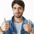 Close-up shot of satisfied and happy young attractive male with blue eyes and bristle in denim jacket raising thumbs up and smiling cheerfully as being pleased with good quality of clothes he bought.