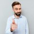 Sly smiling bearded man in business clothes pointing and looking at the camera over gray background